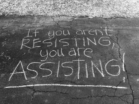 The image shows a message written in white chalk on a cracked asphalt surface. The text reads:
“If you aren’t RESISTING, you are ASSISTING!”
The words “RESISTING” and “ASSISTING” are written in all capital letters for emphasis. The background includes a patch of dry grass at the top of the image. The overall tone is one of activism or protest.