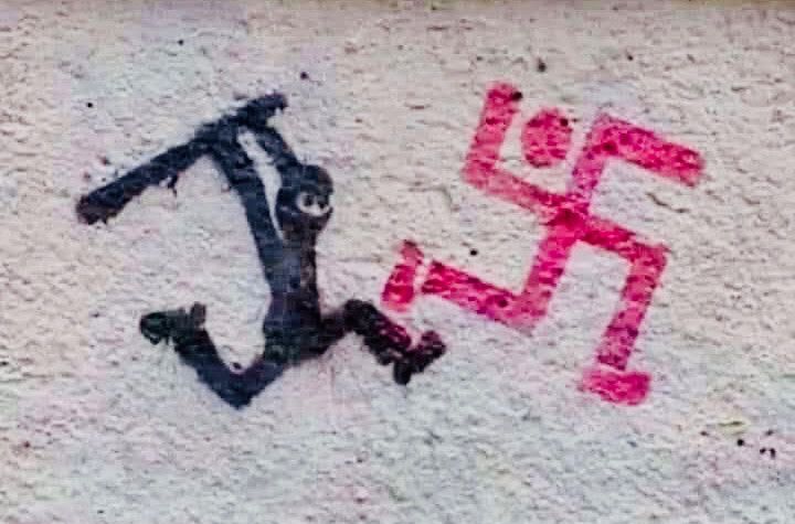 The image is a piece of street art painted on a wall. It depicts a black figure resembling a resistance fighter or partisan, possibly holding a weapon, kicking a red swastika-shaped figure that has human-like arms and legs. The swastika figure appears to be falling or retreating, emphasizing the idea of resistance against fascism and Nazism.

The artwork symbolizes opposition to fascism, suggesting that those who stand against it will fight back and ultimately defeat it. The simple yet powerful imagery conveys a strong anti-Nazi and antifascist message.