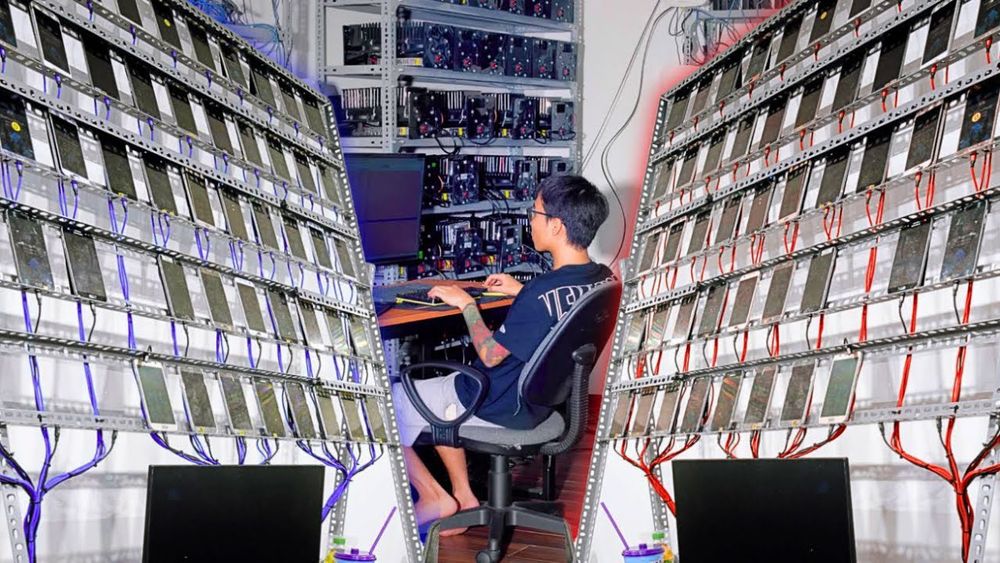 A young man with short dark hair sits on a black office chair at a desk in a high-tech setup resembling a troll farm, typing on a keyboard in front of a computer monitor. He wears a dark blue t-shirt and light-colored pants, with one foot casually extended. Flanking him on both sides are tall, angled racks densely packed with hundreds of mounted smartphones or tablets in black cases, connected by a web of red and blue cables dangling from above. The composition forms a V-shape around him, emphasizing the overwhelming scale of the operation, with dim lighting and additional server equipment visible in the background.