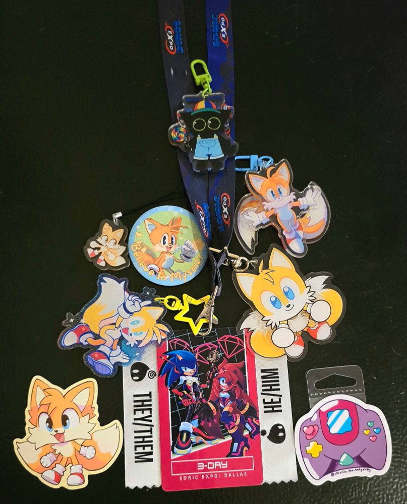 A collection of the merch I got from the convention. It is array of keychains of Tails, a sticker of a Dreamcast controller, and the badge from the convention. On the badge are two pronoun ribbons- one with He/Him, and the other with They/Them.