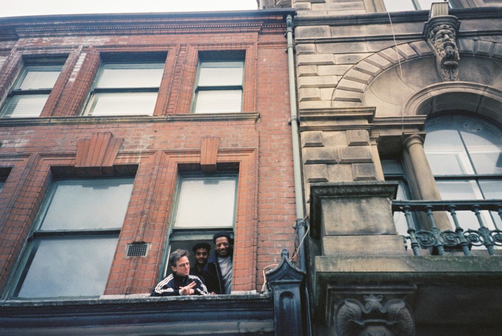 Three people look out of the SADACCA building window. The building is red brick with black window frames.
