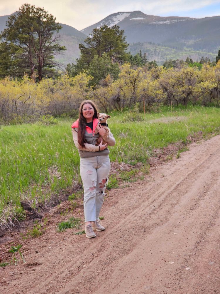 Leah, a woman with reddish/brown hair and blue eyes, is smiling while holding a small brown dog. Behind her is a field full of grass, bushes, and trees all of which are green. In the background are mountains.