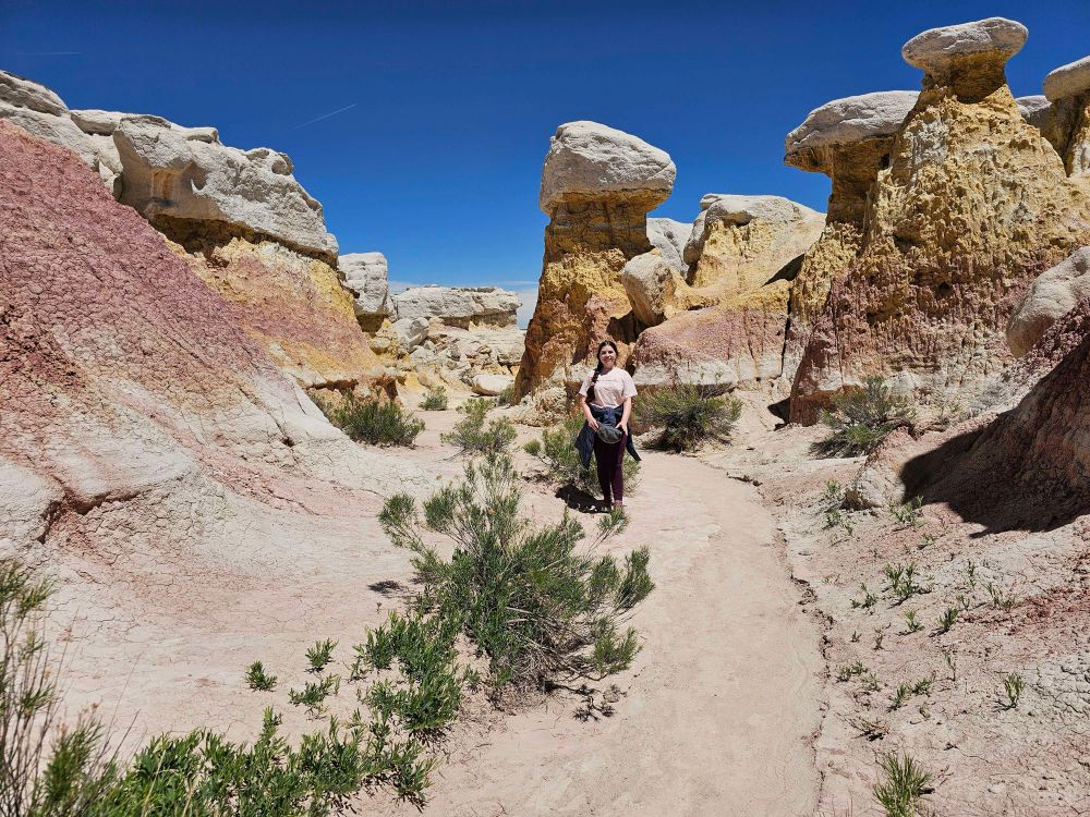 A woman is standing on a sandy trail surrounded by tall, multicolored rock formations in a desert canyon. The rocks display layers of white, yellow, and pink hues under a bright blue sky.