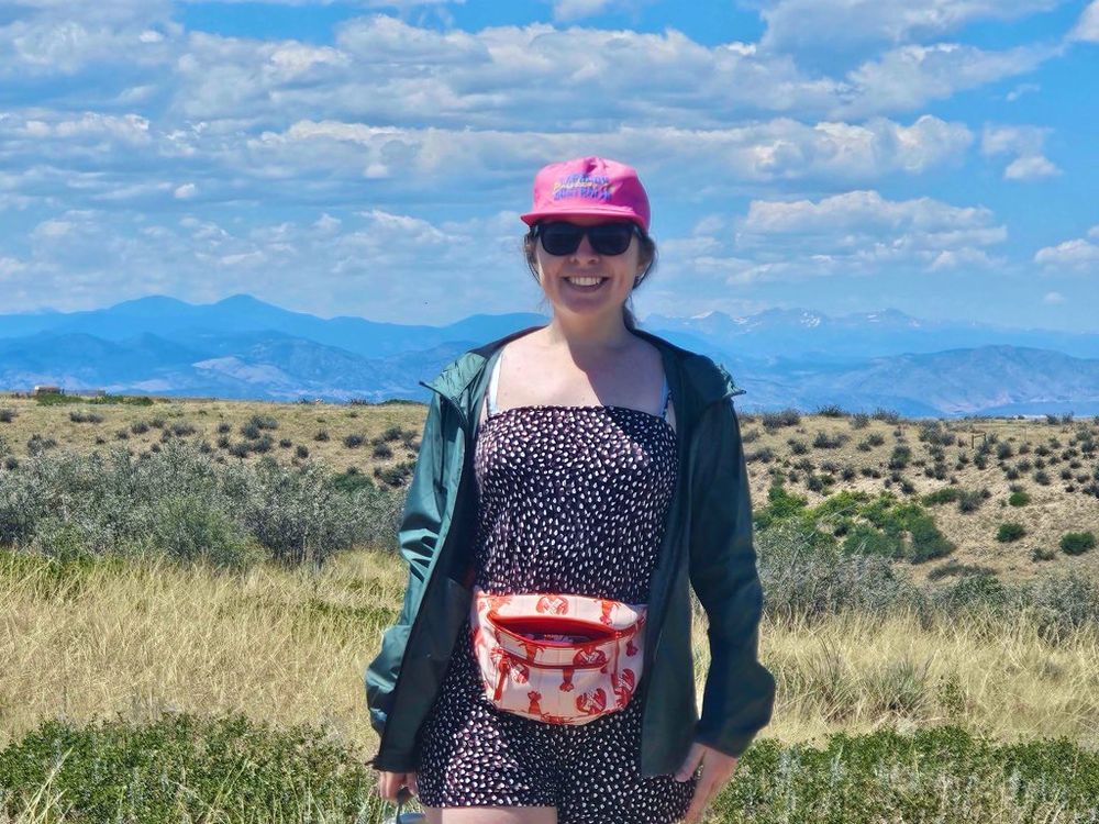 Leah, a woman with reddish/brown hair in a ponytail and blue eyes, is smiling towards the camera. Behind her is an open field and further back are the Rocky Mountains. Leah is wearing a pink hat that says “Laracon AU Brisbane ‘24” on it.