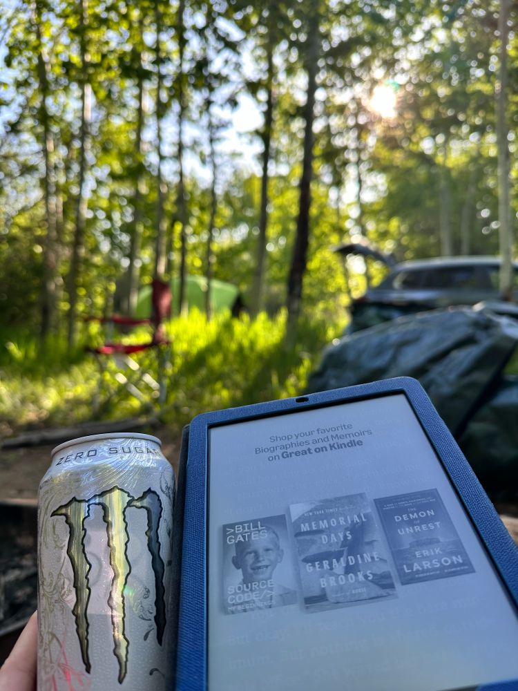 Leah is holding a white monster while a Kindle Paperwhite is sitting to the right of the monster. In the background is a tarp lying on the ground and a red camp chair to the left of the tarp. In the background are a green tent and a blue/gray Mazda CX-50 which has its trunk open.