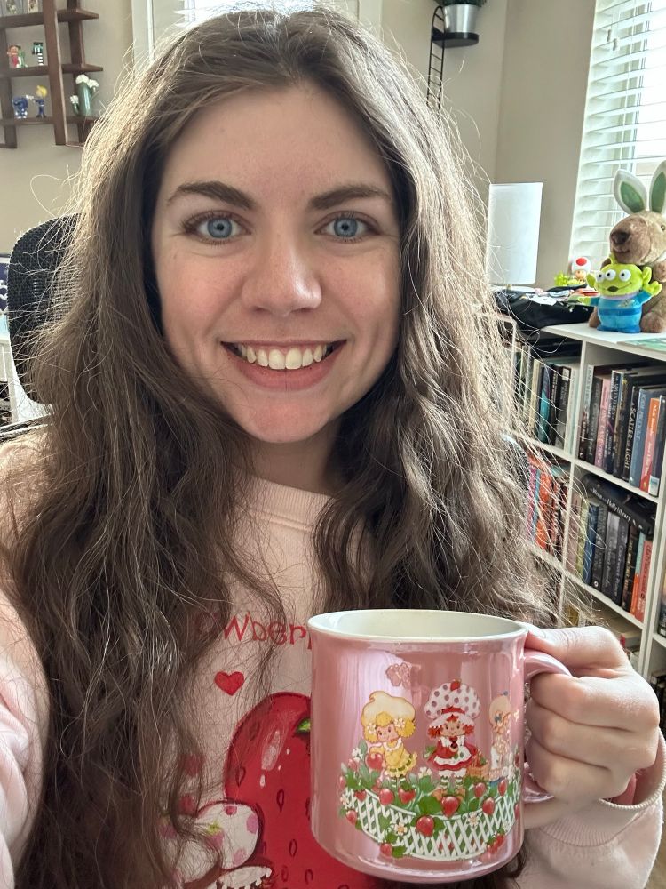 Leah, a woman with wavy reddish/brown hair and blue eyes, is smiling towards the camera while holding a strawberry shortcake mug full of coffee