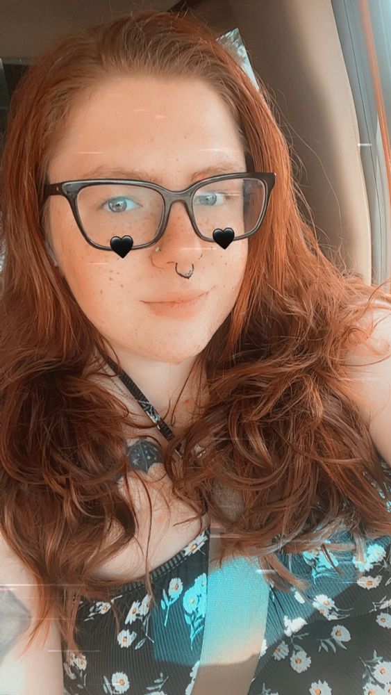 A selfie of a woman with red hair and black glasses wearing a black sundress with daisies on it