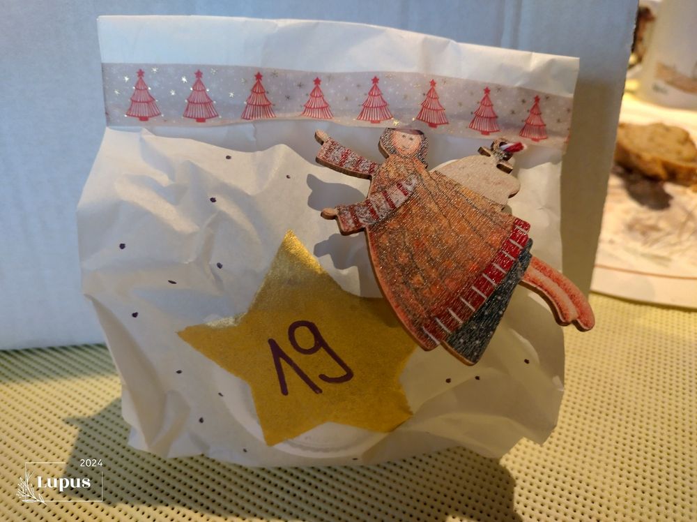 Eine weisse Tüte mit den Holzengeln. Ein goldener Stern mit einer 19.
Zugebunden mit einem roten Band mit weissem Tannenbaum.


