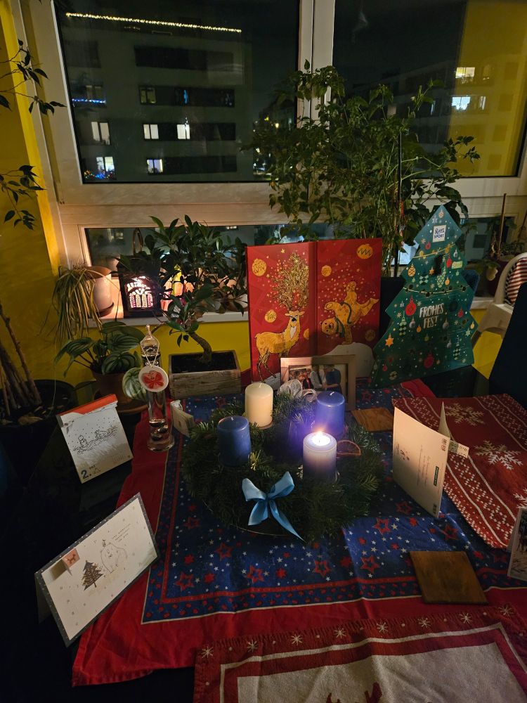 Our dining table (black and square, with a glass top). There is a red, blue and white table cloth with snowflake patterns in the middle, and two paper Advent calendars can be seen in front. Two red and white placemats are on top of the tablecloth. At the centre, there is a new Advent wreath with three blue candles and one white candle. The bottom right blue candle has been lit. Two chocolate Advent calendars are propped up against a chair.  In the back,  you can see the bright yellow wall, and the window with several plants and a wooden lantern with coloured paper motifs  (looking like a church window). A giant chili plant takes up the middle of the window.