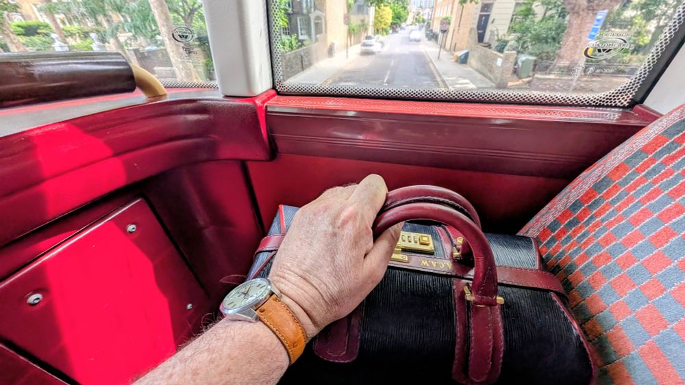 Bright red London bus interior with red moquette seats, view out window, trees and red brick houses typical London. Hand holding an elegant red and black leather doctors bag