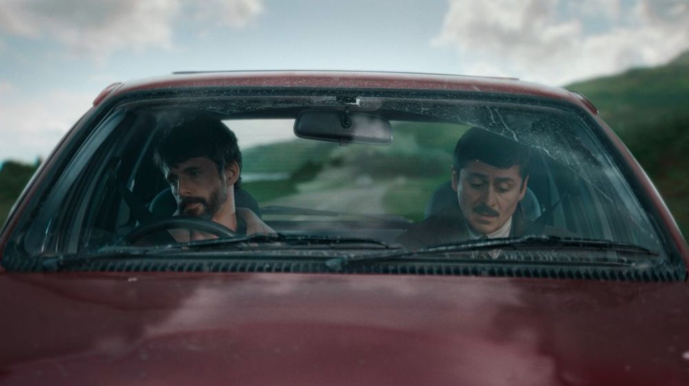 Akram and Morck pretending to drive a C reg Sierra in a studio with a green screen behind them showing the Scottish countryside. 