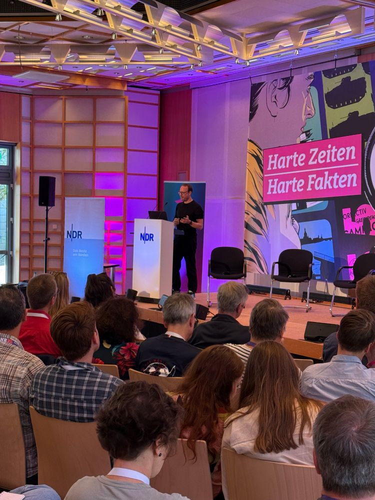 Der Journalist Markus Beckedahl hält seine Keynote-Rede hinter einem weißen Pult mit der Aufschrift „NDR“. Er steht auf einer Bühne. Dahinter hängt ein Banner mit comicartigen Nachrichten-Motiven und der Aufschrift „Harte Zeiten, Harte Fakten“. Vor der Bühne sitzen Teilnehmende der Netzwerk-Recherche-Jahreskonferenz. 