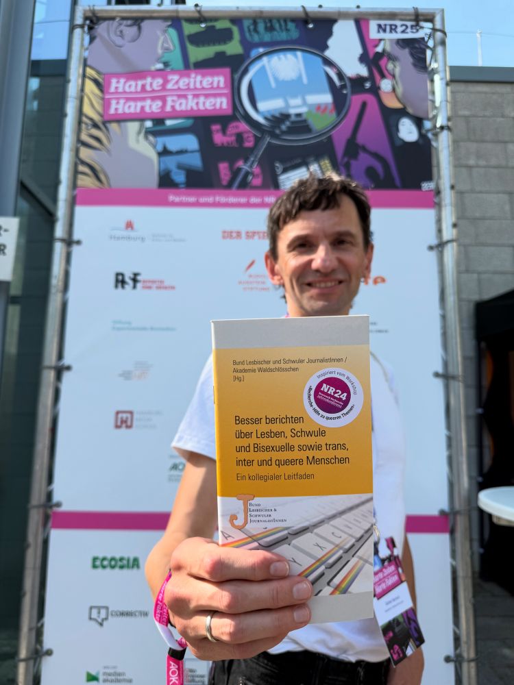 Ein Mann mit weißem Shirt hält die „Besser berichten“-Broschüre in Richtung Kamera. Im Hintergrund ein Banner der NR-Jahreskonferenz.
