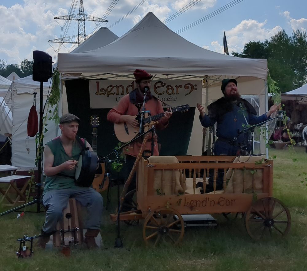 ein weißer mittelalterlicher Pavillon unter dem ein Barde mit Gitarre musiziert, ein sitzender Mann Bodhrán spielt und ein bärtiger Mann fürs Entertainment sorgt