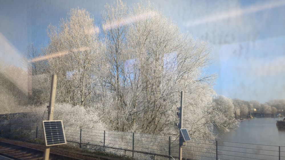 Durch die Spiegelung eines Zugfensters fotografiert: 
Gefrorener Fluss unter einem Bahnübergang, schneeweiße Bäume dahinter. Die Bäume sind primär mir Raureif bedeckt, der wie Schnee anmutet.