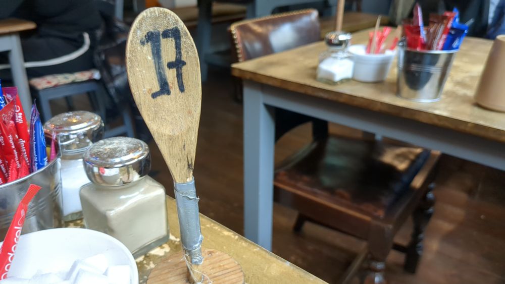 ein abgewetzter Holztisch in einem urigen Restaurant. Auf dem Tisch stehen Gefäße mit Pfeffer und Salz und Eimerchen mit Ketchup, Senf, Brown Sauce. Ein abgebrochener Löffel, der mit Panzertape auf eine kleine Holzplatte geklebt wurde zeigt eine schwarze 17. 