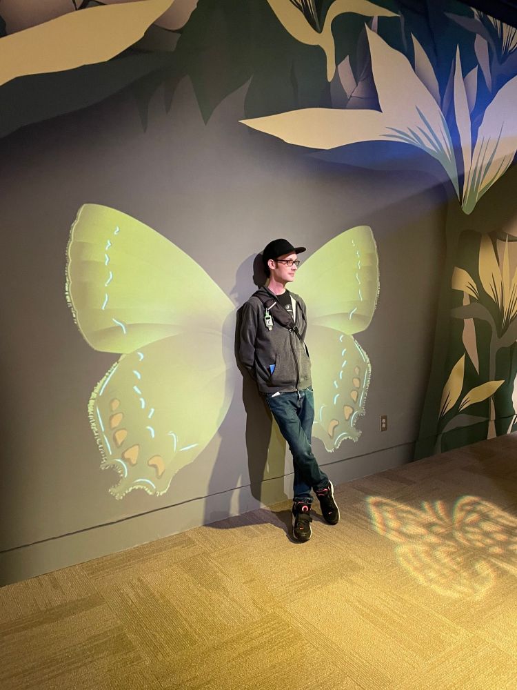 Wesley is standing cross-legged in front of butterfly wings on the wall.