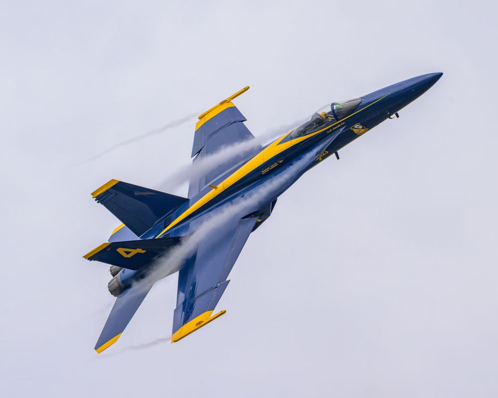 Blue Angels jet #4. The navy blue plane with yellow striping passes over the photographer, right wing down. The pilot is visible through the canopy. Vapor trails form below the canopy and extend over the wings. 