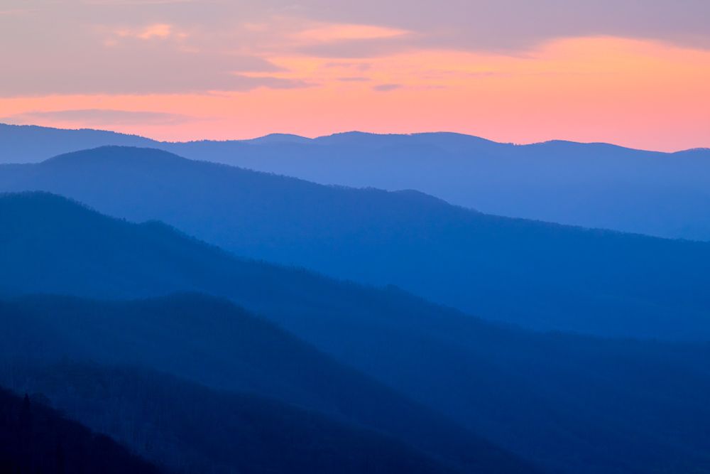 Silhouettes of mountain ridges extend towards the horizon. The haze in the mountains appears blue. Above the horizon is pink sky, with clouds above.