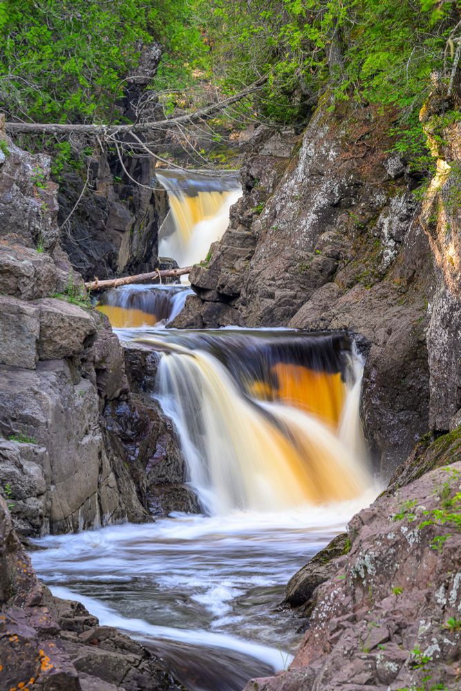 A series of short waterfalls descending through a granite canyon. Lush green foliage can be seen at the top of the photo. The water is stained brown from the peat bogs where the river originates. 