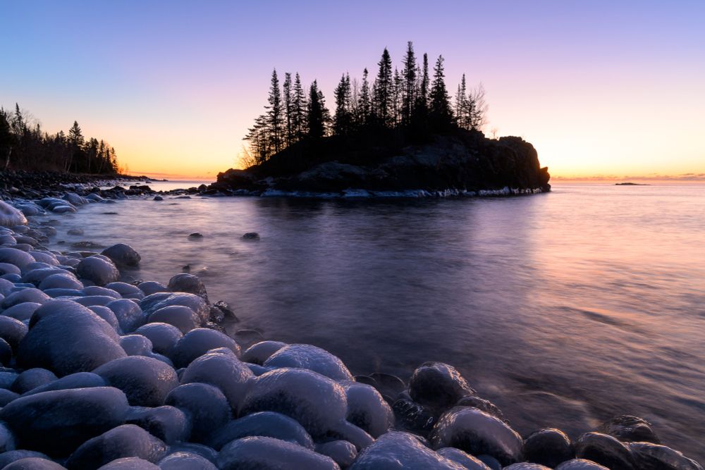 A winter scene along Lake Superior, taken 25 minutes before sunrise. The scene is lit in a split color tone. The shadows are a cool purple color typical of early dawn before sunrise. The highlights are a pale golden color.

In the foreground, extending from the bottom right to the middle left, are icy boulders. The ice appears pale purple, with the sunward-facing portions lit in pale gold. Beyond the boulders, pine trees grow along the shore. In the center of the image is a rocky island, connected to the shore by a narrow strip of boulders. The island appears mostly dark as the sun is rising behind and to the right of it.