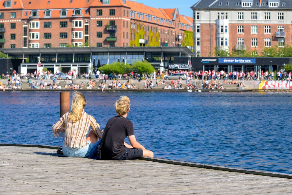 A young couple sits on a wooden walkway along a river in the sun. Across the river, many people are seated on concrete benches along the river.