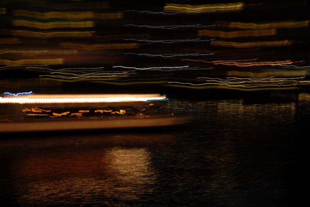 A long-exposure shot of a boat moving along a canal at night. The camera has been panned faster than the speed of the boat, causing the boat and lights on buildings behind the boat to be blurred into colorful lines. The boat is not so blurred that it is unrecognizable.