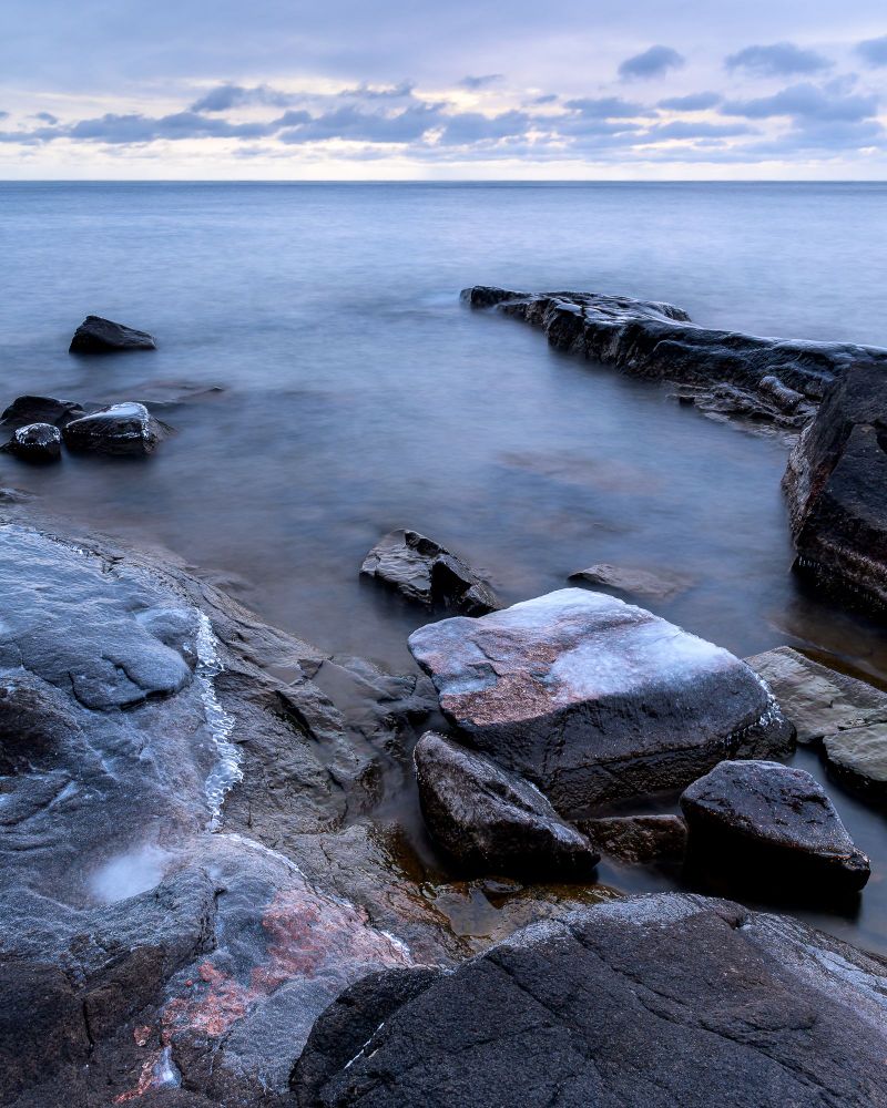A scene along Lake Superior. The general feeling of the photo is flat and empty.  The sky is gray, with dark lower level clouds beneath a higher, flat gray ceiling. Calm waters and long exposure have made the water smooth and flat.  Black basalt, interspersed with red granite, and covered in ice, surround a small bay filled with boulders. On the back side of the bay, a long spit of black basalt points out toward the horizon.