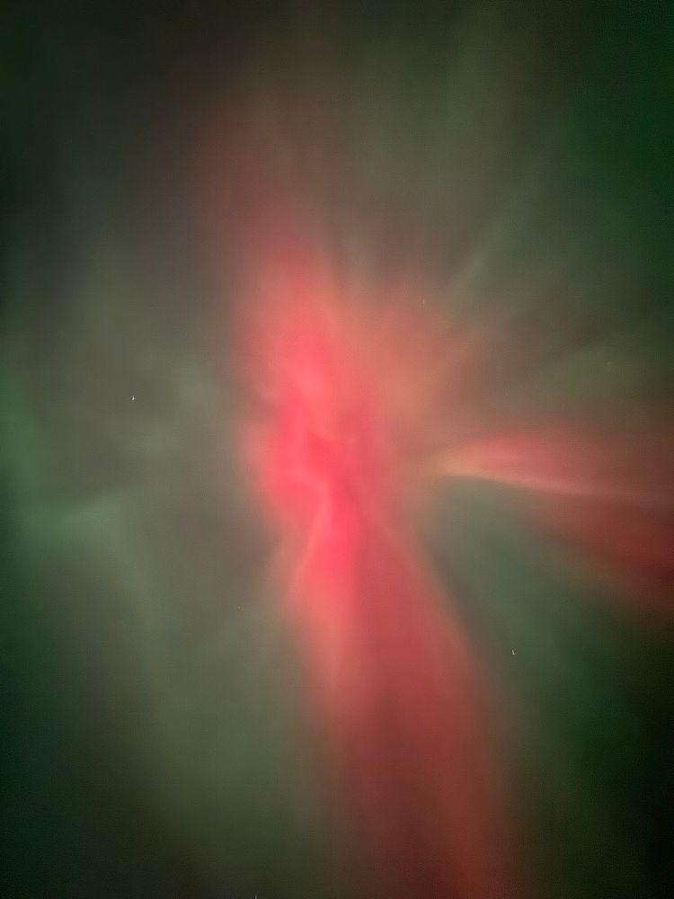 A photo of the northern lights. A bright red beam runs vertically through the center of the image, surrounded by dimmer green and dark sky
