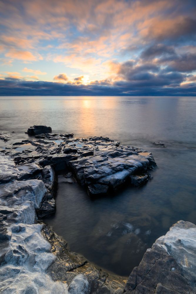A sunrise scene over Lake Superior. The foreground is comprised of black basalt, partially covered in ice. There is a j-shaped channel worn in the basalt, with the top of the j angled 45 degrees to the right so that the inner point of the j points toward the horizon where the sun is coming up. The lake is calm, allowing the light in the sky to be reflected on the lake's surface. On the horizon, dark, heavy cloud cover gives way to partially cloudy skies filled with thin, puffy clouds. Golden light from the sun appears in the center above the clouds, illuminating the fluffy clouds in a pale orange and peach light.