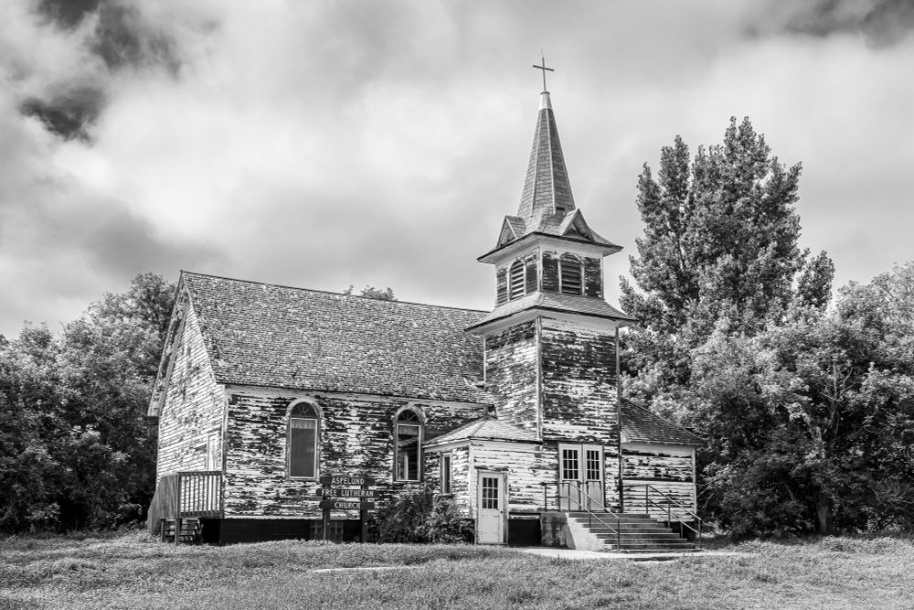 A black and white photo of an old prairie Lutheran Church sits in a grassy clearing amongst trees. The church is built in the typical style of these churches - a rectangular building with an entryway built onto the side. The bell tower sits above the entryway. The building is shingled with wood shakes. The sky above the church looks moody with very dark areas (actually blue sky in the color version), with darker and lighter clouds.