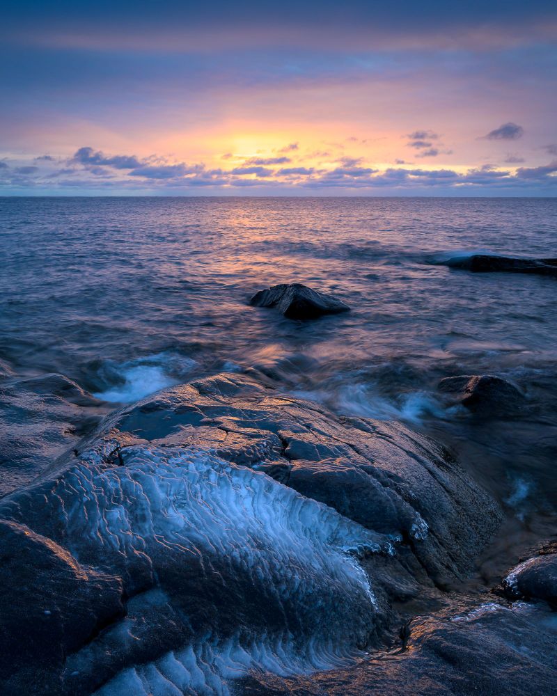 A cloudy sunrise scene over Lake Superior. The sky is lit in a peach color, with a golden yellow orb near the sun which is behind the clouds. Low clouds over the lake are a darker, almost pale purple color. In the foreground, a black basalt formation that is roughly triangular extends into the lake. In the immediate foreground, ice covers the basalt, ranging from clear ice reflecting the color of the sky to thicker white ice. The thicker ice is ridged and the edges of the ridges also reflect peach color in the sky.