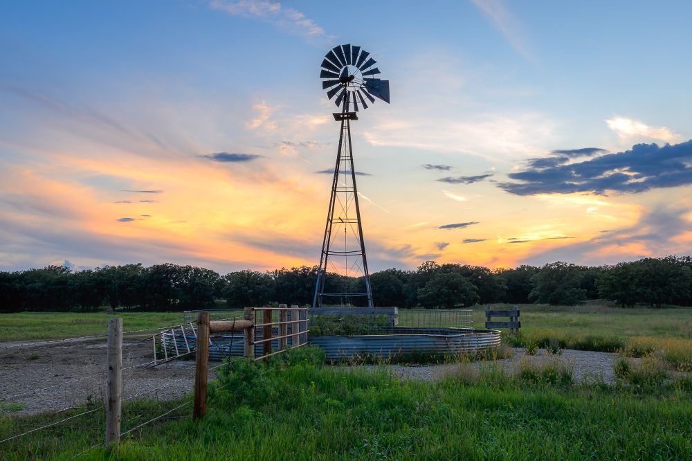 A sunset scene. In the foreground, a barbed wire fence extends from the bottom left towards a windmill and stock tank in the center. Lush green grasses fill a meadow. At the back of the meadow, oak trees grow on gently sloping hills. The sky is partly cloudy. The sun, already below the horizon, lights wispy higher clouds in pale orange and yellow, while lower clouds appear darker and blue.