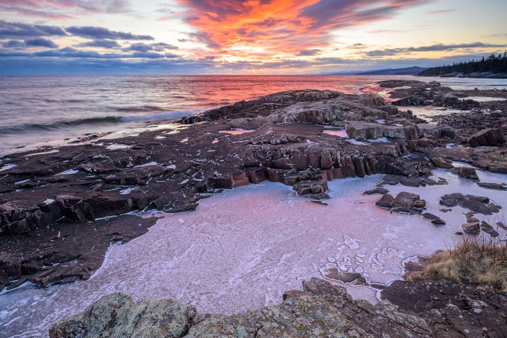 A vivid sunset scene along Lake Superior. In the center top of the photo, is a dark, wedge-shaped cloud. The underside of the sky is lit in in vivid orange and pink by the sun which is below the horizon. The unlit parts of the cloud are darker purple.  The water beneath the cloud is red, reflecting the color of the sky.

In the foreground is red granite rock, covered in gray lichens. Beyond that is a sheet. The ice reflects the pink hues of the sky. Beyond the ice is a maroon rock formation. Spots of ice on the rock reflect the vivid hues of the sky.
