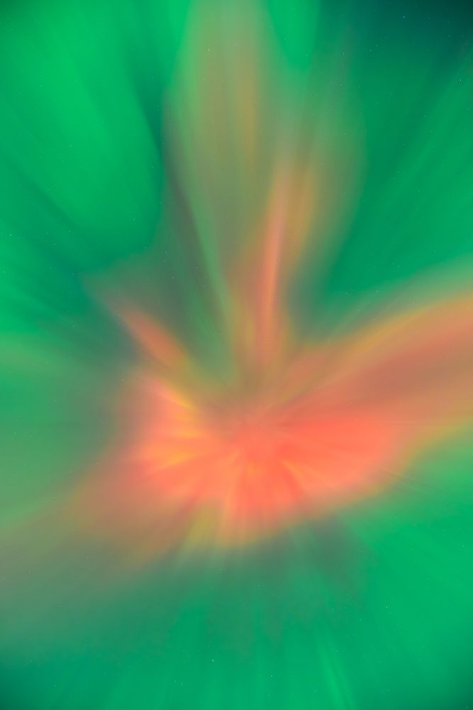 I'm not even sure how to describe this. The photo was taken by pointing the camera straight up in the sky to capture the aurora corona. Slightly below the center of this vertical image is a red shape, like a flattened ball, with two lobes pointing upwards. Yellow and orange highlights appear in the red. The red is surrounded by a greenish blue cloud of aurora.  The aurora is so bright, the stars above are only slightly visible by zooming in on the picture. The stars appear as dim white dots behind the aurora.