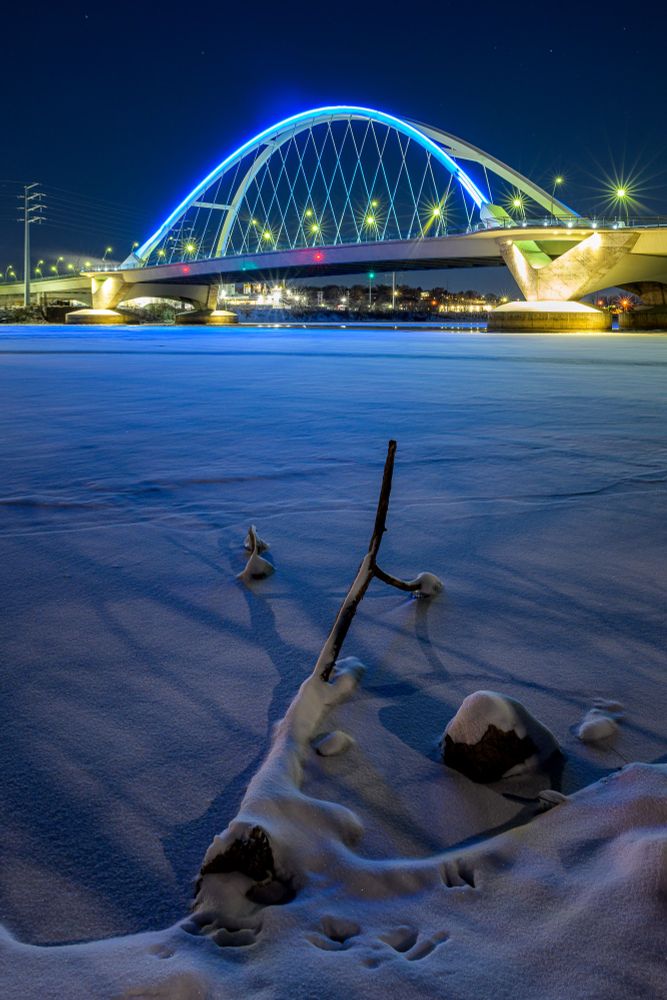 A vertical image of a winter scene at night. The Lowry Ave. Bridge spans the frozen Mississippi river. The center of the bridge features an arch with support poles that appear as a crosshatch pattern. The arch of the bridge is lit with a blue light. In the foreground, snow covered rocks and a fallen tree limb are covered with snow. The tree limb extends from the lower right corner, pointing towards the arch of the bridge.