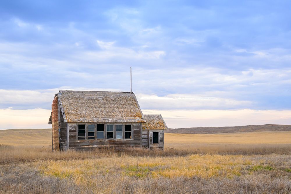 A small one-room schoolhouse on the plains. The school is viewed from the rear corner of the school. A brick chimney is still standing but is separated from the building, leaning slightly away from the building. There are six windows on the side of the school. Some of the windows are broken. The door in the entryway is closed but the window there is broken. A flagpole extends above the building. The roof is mostly intact, covered in wooden shake shingles, with only a few small holes.

Surrounding the building are dried brown and gold prairie grass. Further behind the school is the stubble of a harvested wheat field covering rolling hills. Further behind is a grass-covered hill. The sky above is filled with clouds that appear blue and gray.