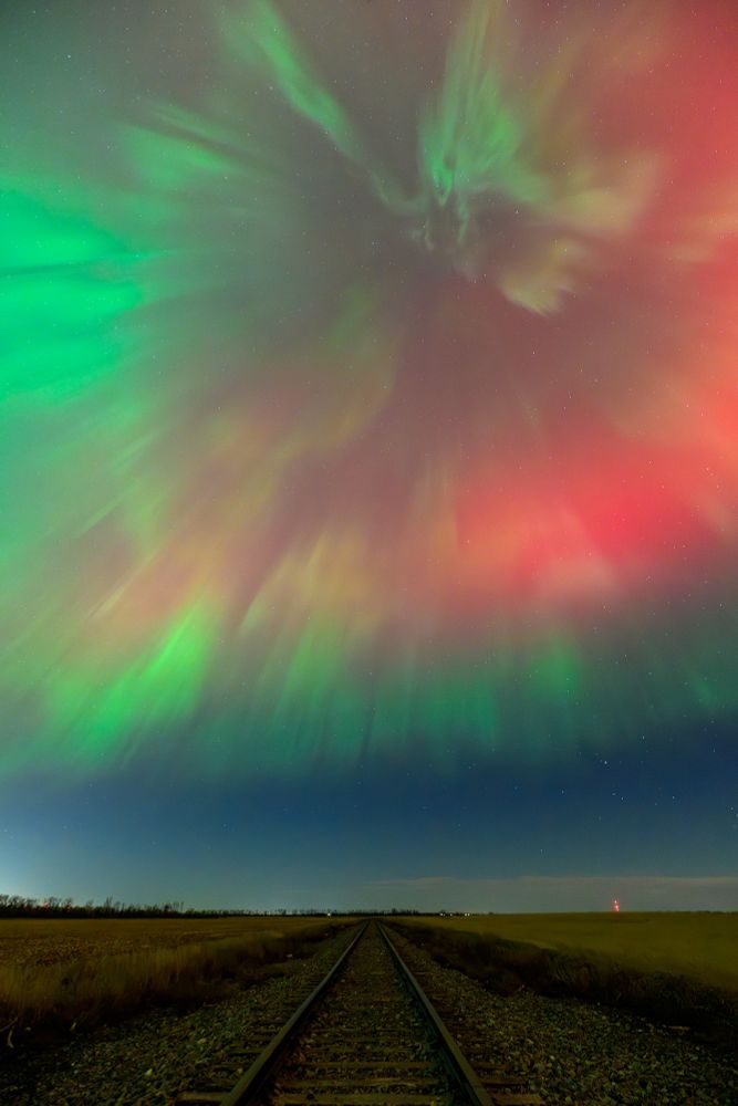 Train tracks extend to the horizon beneath an auroral corona. The sky on the horizon is dark blue. Above, the corona shows as ribbons of green light, with a red glow extending from the upper right to the lower left side of the corona.