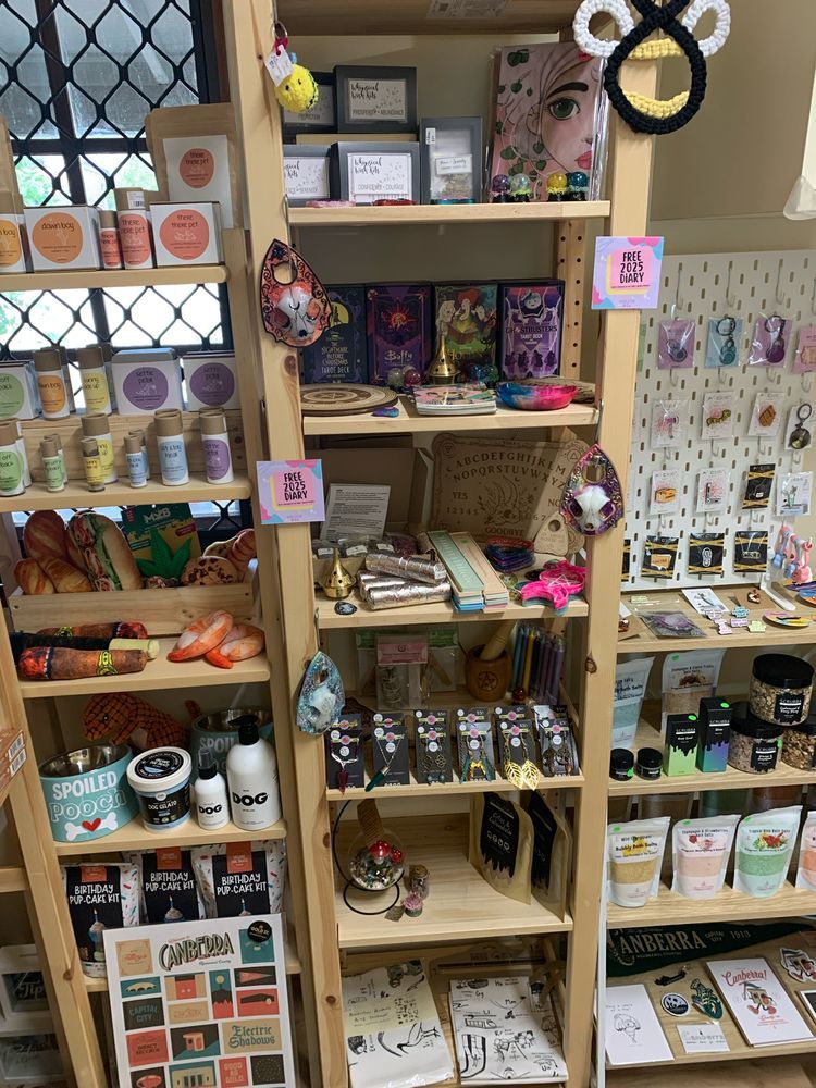 Image shows light wooden shelving with various items on the shelving from different artists and small businesses. 

From Attire Dolls the image shows large cat skull planchettes hanging from hooks, a crescent moon trinket dish, two triple moon trinket dishes, multiple different coloured resin crystal balls, and other sized planchettes. 

Standing up on one of the shelving against dark backing cards is some glasses chains and necklaces, and on the bottom shelf are some crystal terrariums. 