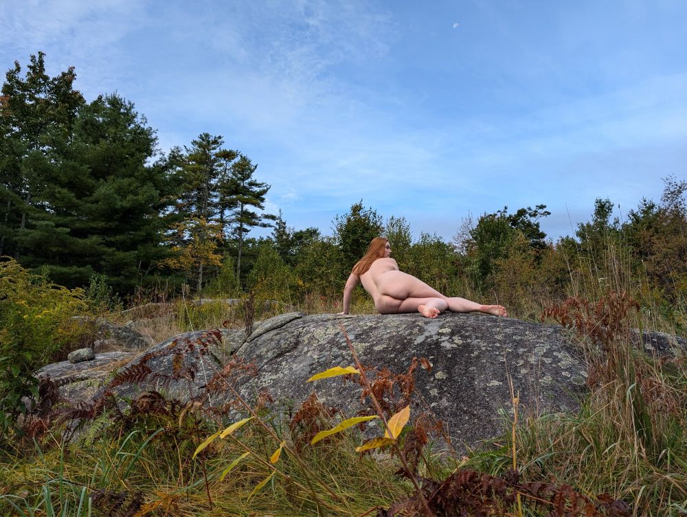 Plus size woman laying on a large boulder in a field during the day. Her naked backside is facing the camera. The woods are behind her and the half moon is in the blue sky above.