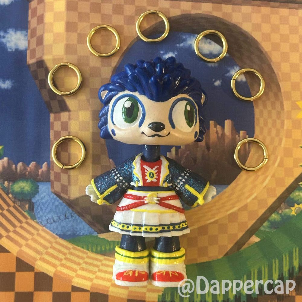 A handprinted custom hybrid mini doll inspired by Sonic the Hedgehog, standing in the green hill zone, surrounded by little golden rings. 

Head came from a Littlest Pet Shop Hedgehog and the outfit/body came from a Yummiland keychain doll.