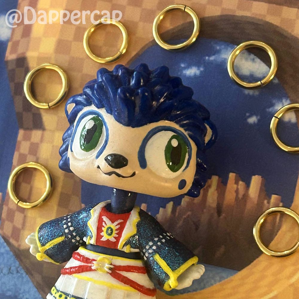 A handprinted custom hybrid mini doll inspired by Sonic the Hedgehog, standing in the green hill zone, surrounded by little golden rings. 

Head came from a Littlest Pet Shop Hedgehog and the outfit/body came from a Yummiland keychain doll.