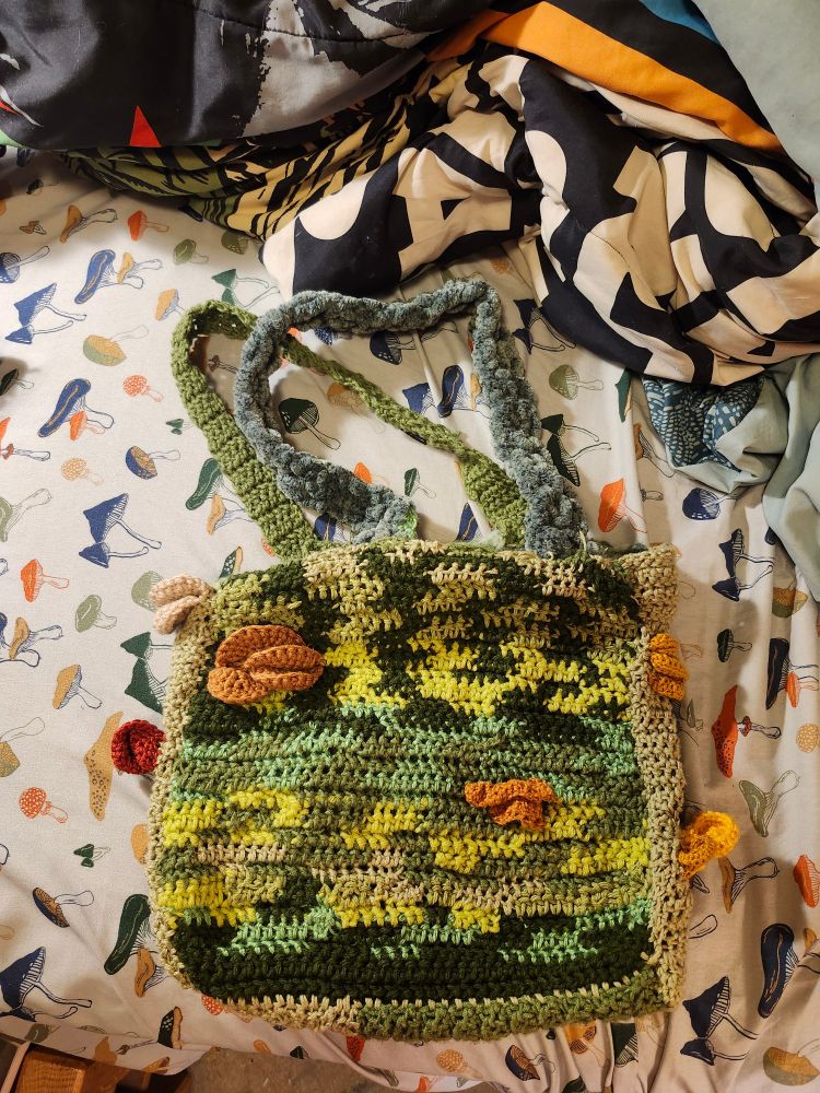 The front side of a crocheted tote bag, the bag is different shades of green and has crocheted mushrooms.