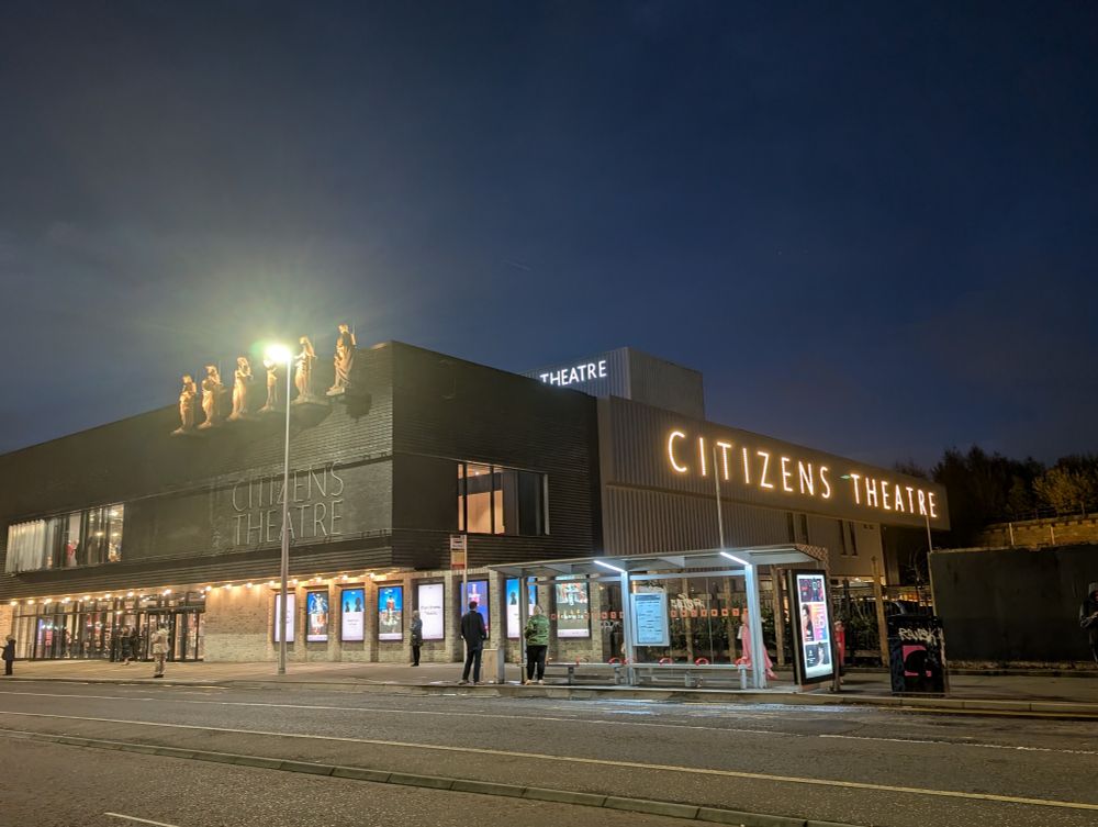 The exterior of the newly restored Citizens' Theatre. The illuminated sign is to the right of the building, the statues of Burns, Shakespeare and the four muses are above and to the left. A bus stop is in front of the building.