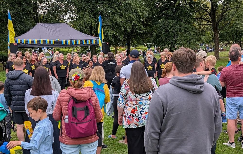 People in the foreground watching a group of singers on a stage in a park, with Ukrainian flags above and to the side.