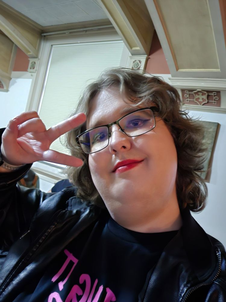 Me, 3 months into HRT at a concert for the Des Moines Gay Men's Chorus. I have shoulder length brown curly hair, with dazzling eyliner and red lipstick. I'm throwing a peace sign and smiling almost smugly at the camera