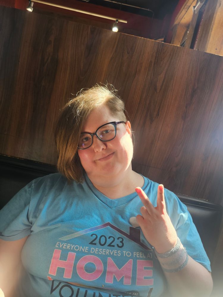 Me, 3 months ago, after I had just gotten my fresh new Undercut haircut. I have rounder glasses now, with my hair appearing more dirty blonde due to the lighting. I am again showing a peace sign.