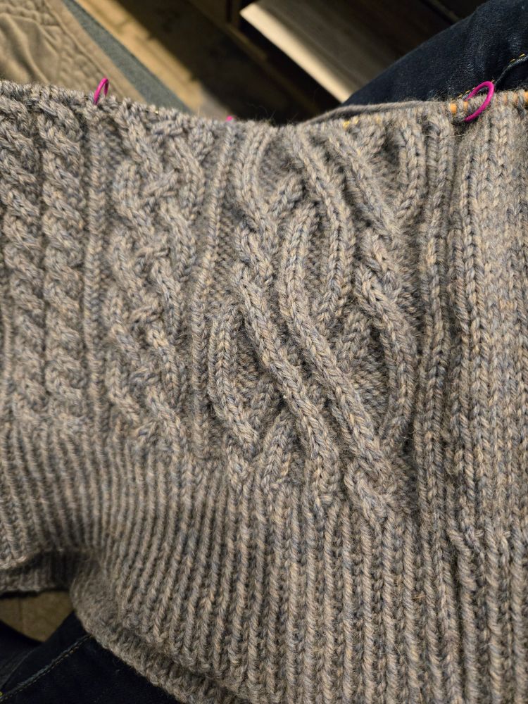 A grey/blue knitting project showing intertwined cable knits as part of the bottom of a sweater 