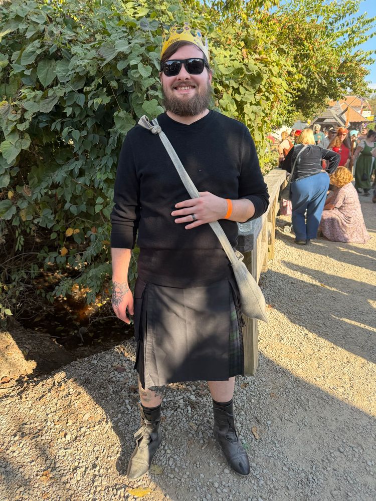 Myself in my day 2 ren faire attire a black thermal shirt, a black utility kilt with green and navy tartan, the same black pointed leather boots, as well as a cotton bag across my body, sunglasses, and a paper crown