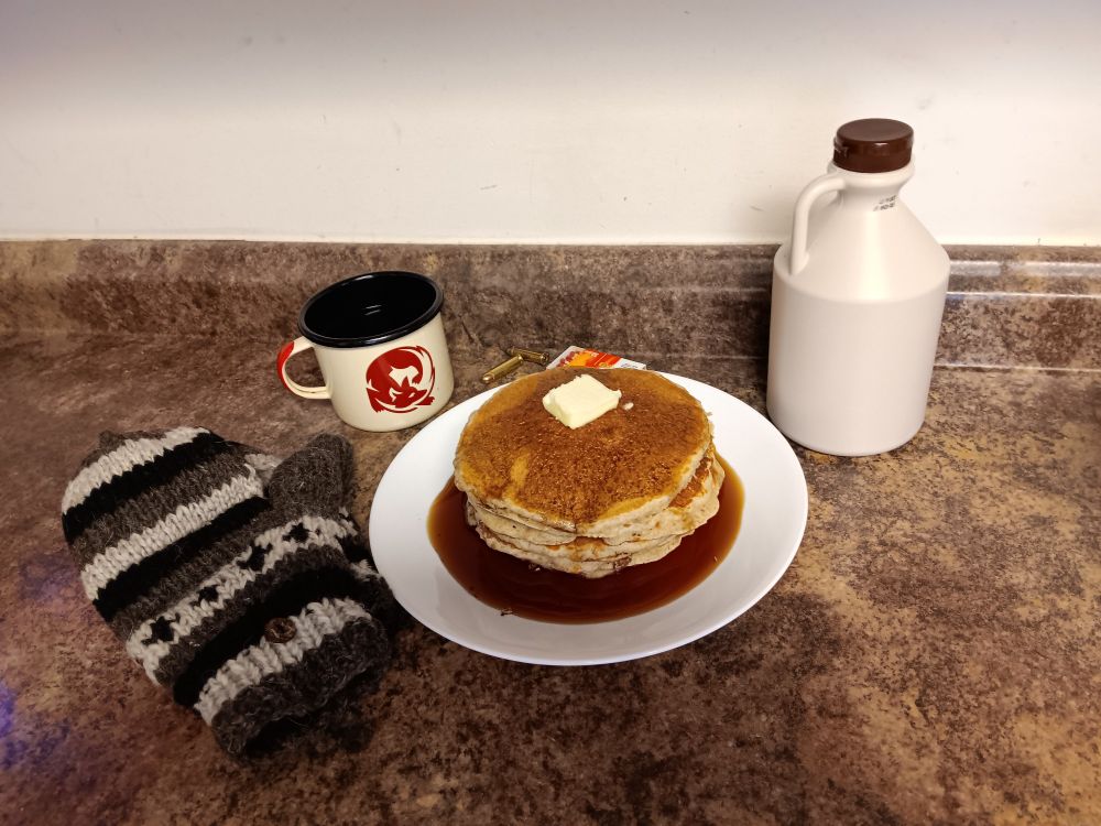 A selection of items similar to game items from The Long Dark. Cup of coffee, wool mittens, stack of pancakes, maple syrup, revolver bullets and wood matches.